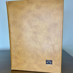 Könnte beinhalten: Ein hellbraunes Portfolio aus Lederimitat mit einer genähten Bordüre. Ein kleines, rechteckiges Emblem mit einem Fahrzeugdesign befindet sich in der unteren rechten Ecke. Das Portfolio steht auf einer Holzoberfläche.