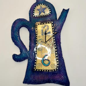 May include: A whimsical, hand-painted wall clock shaped like a teapot. The clock features a blue and purple textured finish, a star design, and a clock face with the numbers 12, 3, and 6. The clock hands are black.