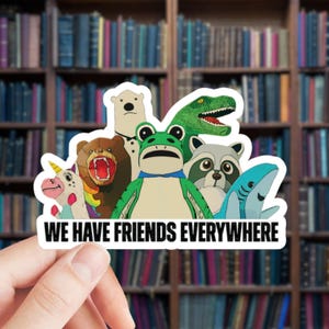 May include: A sticker featuring a group of cartoon animals, including a bear, frog, raccoon, shark, and unicorn. The text "WE HAVE FRIENDS EVERYWHERE" is at the bottom. The sticker is held in front of a bookshelf.