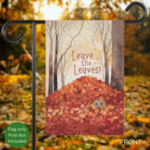 Op de afbeelding: Een tuinvaandel met een aquarel illustratie van een egel in een hoop herfstbladeren. De vlag heeft de tekst "Leave the Leaves!" in een warm lettertype. De achtergrond toont bomen en een herfstkleurenpalet.
