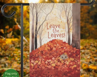May include: A garden flag with a watercolor illustration of a hedgehog in a pile of autumn leaves. The flag has the text "Leave the Leaves!" in a warm, inviting font. The background features trees and a soft, autumnal color palette.