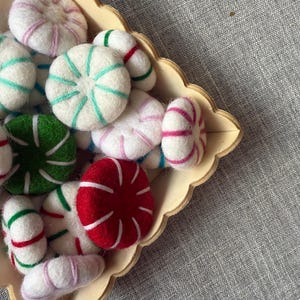 May include: A wooden tray filled with colourful, felted sweets. The sweets are white with stripes of green, pink, red, and blue. Some sweets are round, while others are oblong. The tray has a scalloped edge.