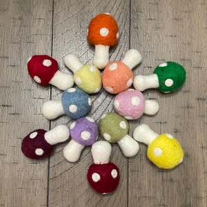 Op de afbeelding: Een verzameling kleurrijke vilten paddenstoelen met witte stelen en witte stippen op de doppen. De paddenstoelen zijn gerangschikt op een houten oppervlak, met kleuren als rood, oranje, geel, groen, blauw en paars.