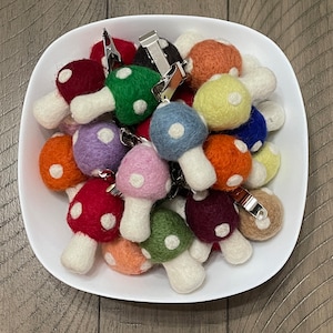 Könnte beinhalten: Eine Sammlung bunter Filz-Pilz-Schlüsselanhänger in einer weißen Schüssel. Die Pilze haben weiße Stiele und Kappen in verschiedenen Farben, darunter Rot, Grün, Blau, Orange und Lila. Jeder Pilz hat weiße Punkte und einen silbernen Clip.