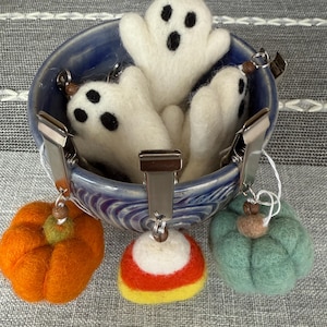 Puede incluir: Un cuenco de cerámica azul lleno de llaveros de fantasmas de fieltro blanco. Clips metálicos están sujetos al cuenco, sosteniendo llaveros de fieltro de calabaza, maíz de caramelo y calabaza verde azulado. Los llaveros son temáticos de Halloween.