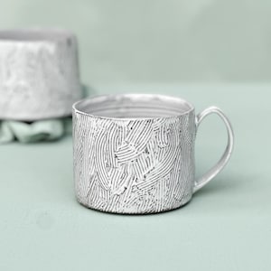 May include: A pair of silver-colored mugs with a textured, lined pattern. One mug is in focus, displaying a handle and the interior. The second mug is slightly out of focus in the background. The mugs are on a light green surface.