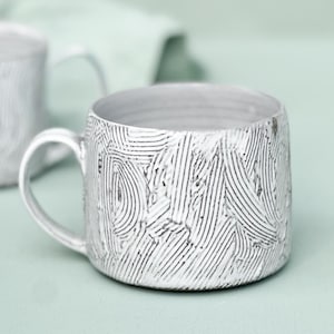 May include: A white ceramic mug with a textured, etched pattern of curved lines. The mug has a rounded body, a handle, and a light-colored interior. The background is a soft green.
