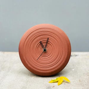 Op de afbeelding: Een terracotta-kleurige, ronde klok met een textuurontwerp van concentrische cirkels. De klok heeft zwarte wijzers en een klein, goudkleurig midden. Een gele bloem ligt bij de klok.