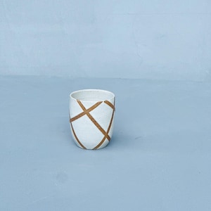 May include: A white ceramic tumbler with a geometric brown crosshatch pattern. The cylindrical cup has a slightly textured surface and a smooth rim. The design is minimalist and modern, suitable for beverages or as a decorative item.