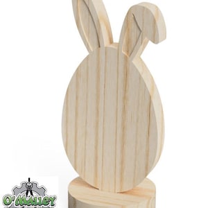 May include: A wooden Easter egg-shaped decoration with bunny ears. The light-colored wood grain is visible. The decoration stands on a round base. The logo "O'Malley Drafting & Designs" is in the lower left corner.