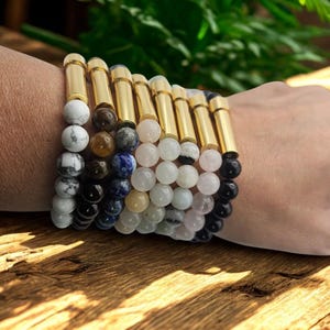 May include: A collection of beaded bracelets with gold-colored cylindrical accents. The bracelets feature a variety of stone beads in colors including white, black, brown, blue, and pink. The bracelets are displayed on a wrist.