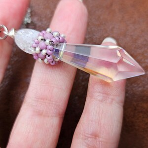 May include: A silver chain necklace with a clear crystal pendant. The pendant is faceted and has a purple and white beaded embellishment at the top.