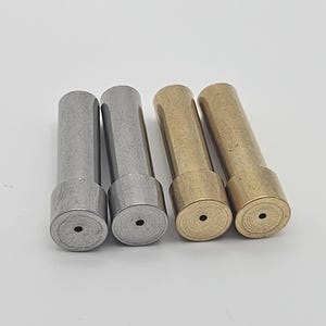 May include: Four metal dowel pins. Two are silver-colored, and two are gold-colored. Each pin has a cylindrical body and a circular base with a small hole in the center. The pins are arranged in a row against a white background.