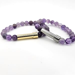 May include: Two amethyst bead bracelets. One bracelet has a gold-colored clasp, and the other has a silver-colored clasp. The beads are a mix of purple and lavender hues. The bracelets are displayed on a white surface.