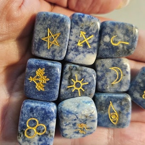 May include: A set of twelve blue lapis lazuli gemstone dice with gold symbols on each side. The symbols include a star, an arrow, a hook, a tree, a sun, a crescent moon, waves, a circle with three dots, a palm tree, and an eye.