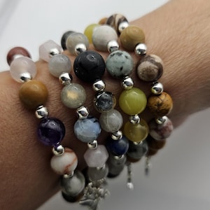 May include: A stack of six bracelets made with various semi-precious stones, including amethyst, labradorite, and jade. The bracelets are strung on silver beads and have a silver charm at the end.