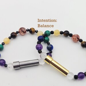 May include: Two beaded bracelets with the text "Intention: Balance". The bracelets feature a variety of colorful gemstone beads, including purple, blue, green, yellow, brown, and clear beads. One bracelet has a silver clasp, and the other has a gold clasp.