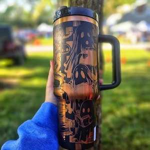 May include: Black and copper travel mug with a handle, featuring a Halloween-themed design. The design includes illustrations of ghosts, bats, and other spooky elements. The mug has a black lid and a copper-coloured band at the top.