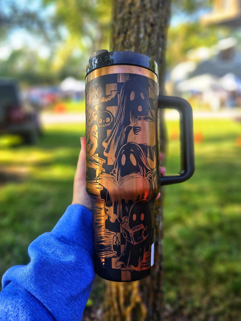 May include: Black and copper travel mug with a handle, featuring a Halloween-themed design. The design includes illustrations of ghosts, bats, and other spooky elements. The mug has a black lid and a copper-coloured band at the top.