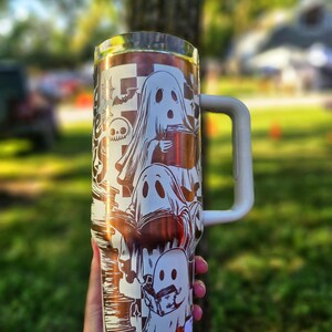 May include: A white and copper-coloured tumbler with a Halloween ghost design. The tumbler has a white handle and a silver rim. The design includes multiple ghosts, bats, and a skull. The tumbler is held outdoors in front of a blurred green background.