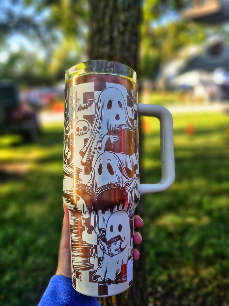 May include: A white and copper-coloured tumbler with a Halloween ghost design. The tumbler has a white handle and a silver rim. The design includes multiple ghosts, bats, and a skull. The tumbler is held outdoors in front of a blurred green background.