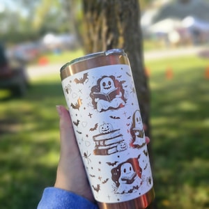 Puede incluir: Un vaso blanco y cobrizo con tem&aacute;tica de Halloween. El vaso est&aacute; decorado con ilustraciones de fantasmas, murci&eacute;lagos, ara&ntilde;as y calabazas. El dise&ntilde;o se repite alrededor del cuerpo del vaso.