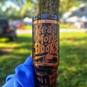 May include: A black and copper-coloured tumbler with the words "Read More Books" in a stylized font. The design includes illustrations of books, a spiderweb, and a ghost. The tumbler has a black lid.