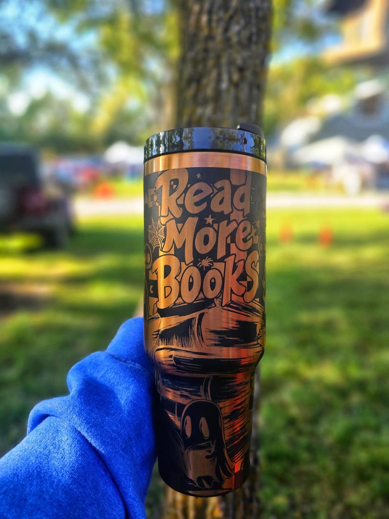 May include: A black and copper-coloured tumbler with the words "Read More Books" in a stylized font. The design includes illustrations of books, a spiderweb, and a ghost. The tumbler has a black lid.