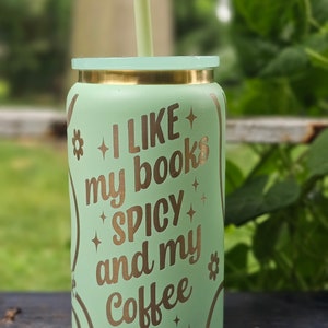 Puede incluir: Un vaso reutilizable verde claro con tapa y pajita doradas. El vaso tiene una inscripci&oacute;n dorada que dice "Me gustan mis libros picantes y mi caf&eacute; helado".
