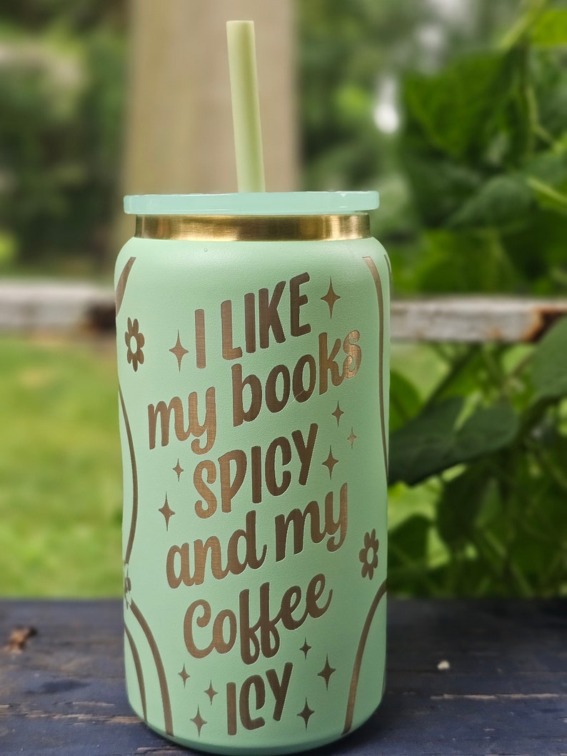 Puede incluir: Un vaso reutilizable verde claro con tapa y pajita doradas. El vaso tiene una inscripci&oacute;n dorada que dice "Me gustan mis libros picantes y mi caf&eacute; helado".