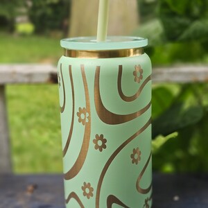 Puede incluir: Un vaso de acero inoxidable verde con tapa dorada y un dise&ntilde;o de remolino dorado con peque&ntilde;os detalles de flores. Una pajita verde est&aacute; en el vaso.