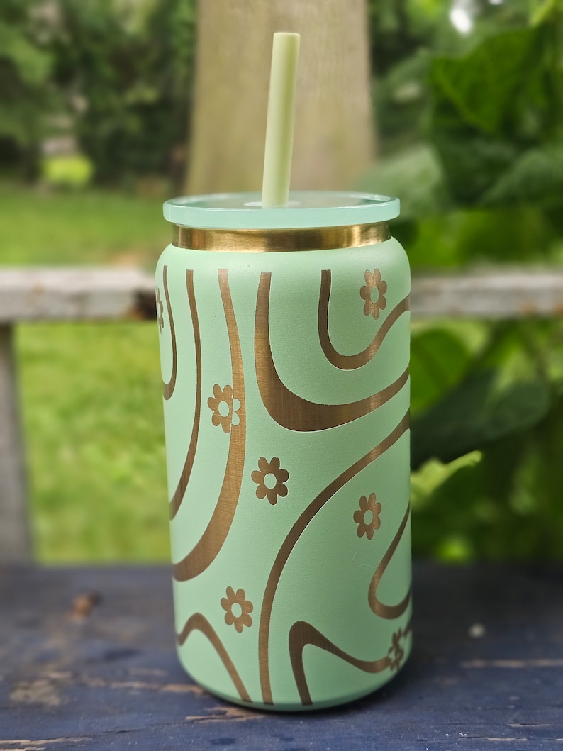 Puede incluir: Un vaso de acero inoxidable verde con tapa dorada y un dise&ntilde;o de remolino dorado con peque&ntilde;os detalles de flores. Una pajita verde est&aacute; en el vaso.