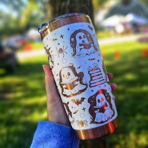 Puede incluir: Un vaso blanco y cobrizo con ilustraciones de Halloween. El dise&ntilde;o incluye fantasmas, murci&eacute;lagos, ara&ntilde;as y libros. El vaso tiene un borde y una tapa de color cobre. Lo sostiene una persona que lleva una sudadera azul.