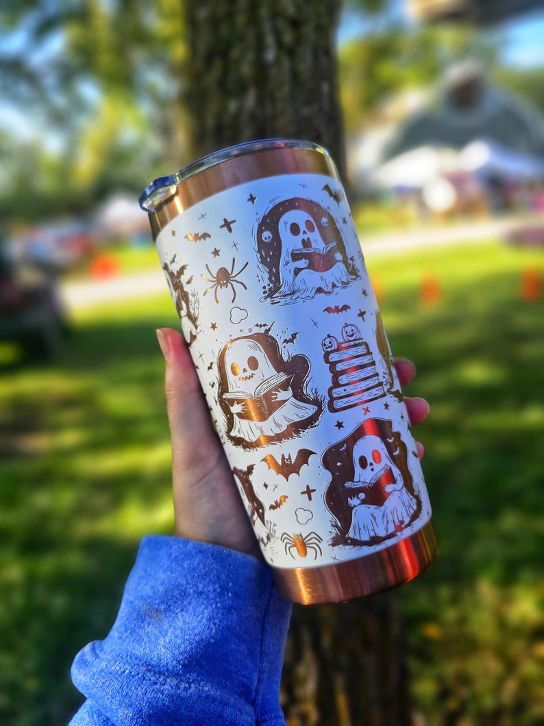 Puede incluir: Un vaso blanco y cobrizo con ilustraciones de Halloween. El dise&ntilde;o incluye fantasmas, murci&eacute;lagos, ara&ntilde;as y libros. El vaso tiene un borde y una tapa de color cobre. Lo sostiene una persona que lleva una sudadera azul.