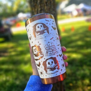 Puede incluir: Un vaso blanco y cobrizo con ilustraciones tem&aacute;ticas de Halloween. El dise&ntilde;o incluye fantasmas, murci&eacute;lagos, ara&ntilde;as y calabazas. El vaso est&aacute; sostenido en la mano de una persona, con un fondo borroso al aire libre.