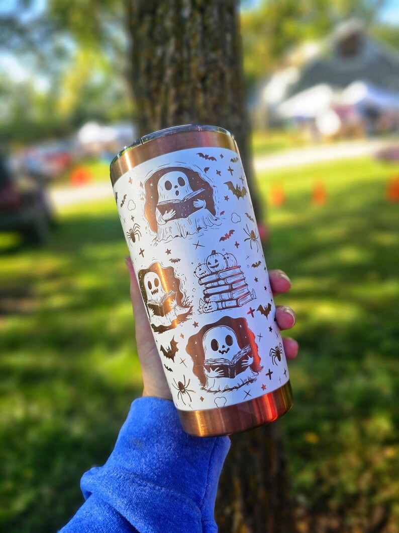 Puede incluir: Un vaso blanco y cobrizo con ilustraciones tem&aacute;ticas de Halloween. El dise&ntilde;o incluye fantasmas, murci&eacute;lagos, ara&ntilde;as y calabazas. El vaso est&aacute; sostenido en la mano de una persona, con un fondo borroso al aire libre.