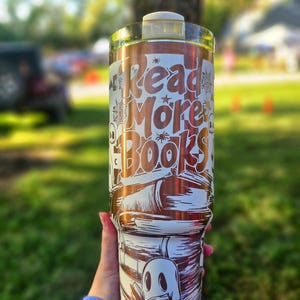 May include: A white and copper-coloured tumbler with the words "Read More Books" in a stylized font. The design includes illustrations of books, spiders, and a ghost. The tumbler is held by a person.