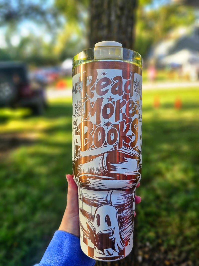 May include: A white and copper-coloured tumbler with the words "Read More Books" in a stylized font. The design includes illustrations of books, spiders, and a ghost. The tumbler is held by a person.