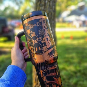 May include: Black and copper-coloured tumbler with a handle, featuring Halloween-themed graphics and the text "Room for Boo". The design includes ghosts, bats, spiderwebs, and a skull. The tumbler is held by a person wearing a blue long-sleeve shirt.