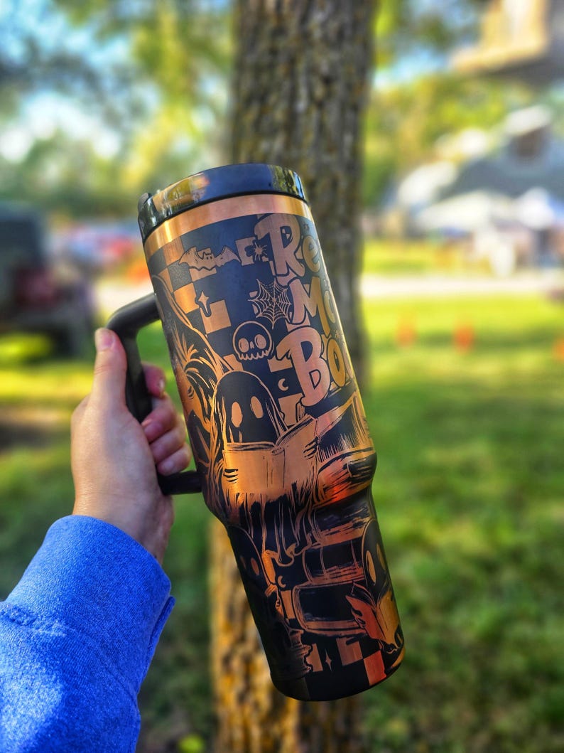 May include: Black and copper-coloured tumbler with a handle, featuring Halloween-themed graphics and the text "Room for Boo". The design includes ghosts, bats, spiderwebs, and a skull. The tumbler is held by a person wearing a blue long-sleeve shirt.