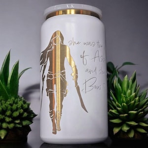 May include: White tumbler with a gold warrior silhouette and gold rim. The tumbler has text that reads "she was the of Ash and she Bow to". The tumbler is surrounded by green plants.