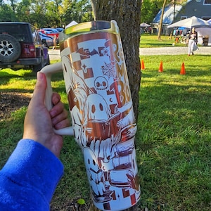 May include: A large, white and copper-coloured tumbler with a handle, featuring a Halloween-themed design. The design includes ghosts, books, and the text "Read More Boo". The tumbler is held in front of a tree.