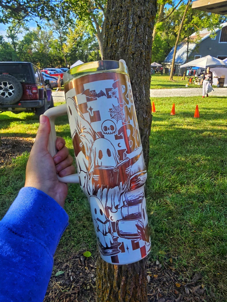 May include: A large, white and copper-coloured tumbler with a handle, featuring a Halloween-themed design. The design includes ghosts, books, and the text "Read More Boo". The tumbler is held in front of a tree.