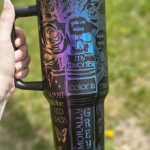 Może przedstawiać: Czarny kubek podróżny ze stali nierdzewnej z tęczowym, opalizującym wykończeniem. Kubek przedstawia czaszkę z różami, motylami i tekstem, który brzmi "you're mine", "my favorite", "color is grey", "I want all the red flags" i "morally grey".