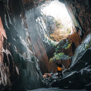 Könnte beinhalten: Eine Person steht in einer großen, dunklen Höhle mit einer kleinen Öffnung oben, die einen hellen, sonnigen Himmel freigibt. Die Höhlenwände bestehen aus rauen, grauen Felsen mit einigen Bereichen aus rötlich-braunem Gestein.