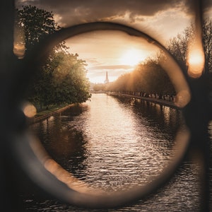 Könnte beinhalten: Blick auf einen Fluss mit einem Kirchturm in der Ferne, gesehen durch eine kreisförmige Öffnung in einem Metallzaun. Die Sonne geht hinter der Kirche unter und taucht die Szene in ein goldenes Licht.