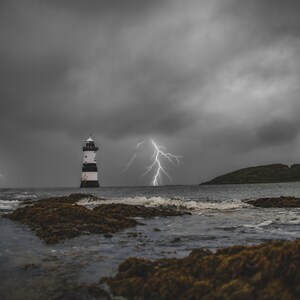 Könnte beinhalten: Ein schwarz-weiß gestreifter Leuchtturm ragt hoch gegen einen stürmischen Himmel mit mehreren Blitzen in der Ferne. Der Leuchtturm befindet sich an einer felsigen Küste, an der Wellen gegen die Felsen schlagen.