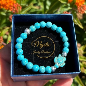 May include: A turquoise blue beaded bracelet with a silver turtle charm and silver beads. The bracelet is in a blue box with the text "Mystic Jewelry Boutique".