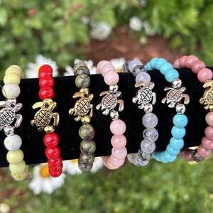 Puede incluir: Siete pulseras de cuentas de colores con un dije de tortuga plateada. Las pulseras se muestran sobre una superficie negra. Las pulseras están hechas con cuentas de diferentes colores, incluyendo amarillo, rojo, verde, rosa, azul y marrón.