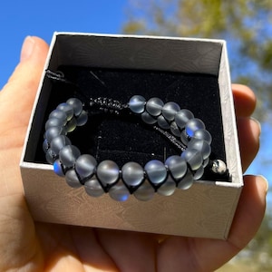 May include: A gray beaded bracelet with a black cord and silver charm in a white box.